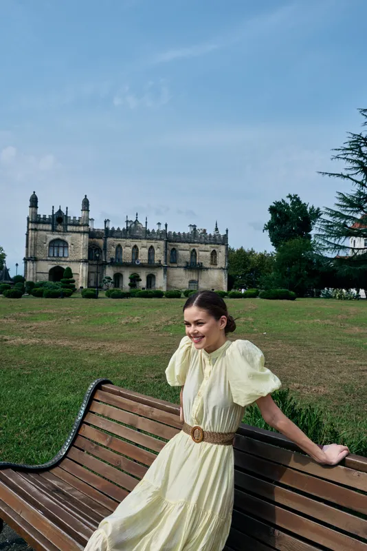 Woman in a pale dress sits on a wooden bench outdoors, with a historic castle in the distance, creating a serene and stylish moment.