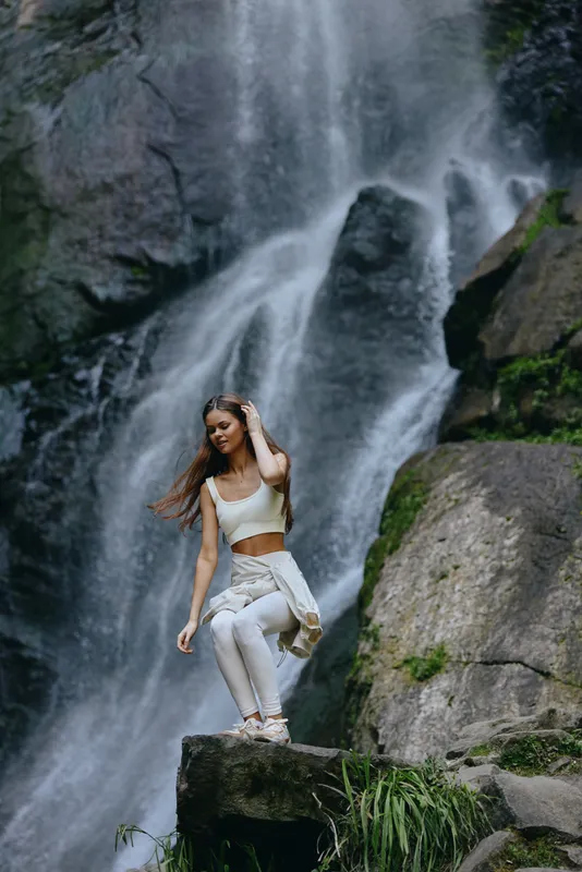 young woman in white sportswear standing on rock near waterfall, nature scenery, outdoor adventure, fitness lifestyle, serene atmosphere, green moss, flowing water
