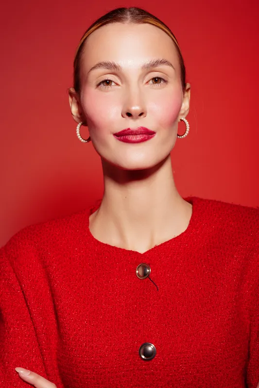 Confident woman with slicked back hair wearing red sweater and hoop earrings poses against red background. Studio portrait with bright lighting and minimal makeup for fashion concept.