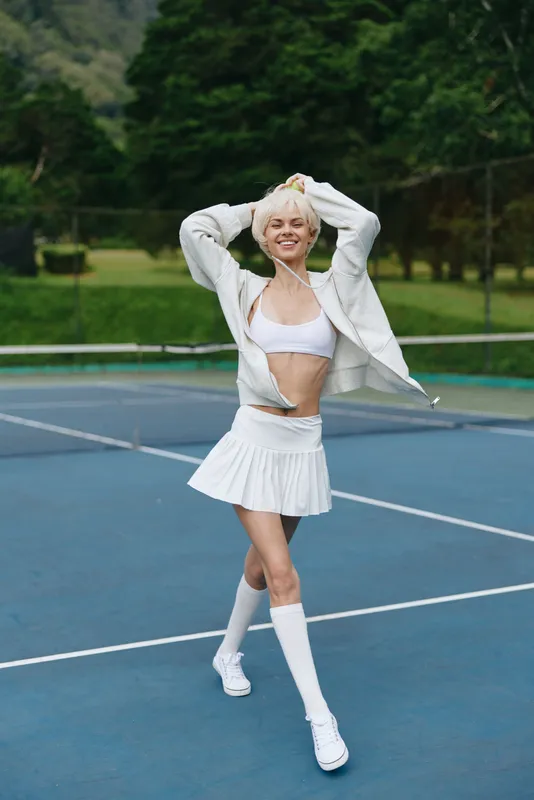 Young athletic woman in white sportswear expressing joy while playing tennis on a court surrounded by lush greenery, showcasing vibrant summer energy