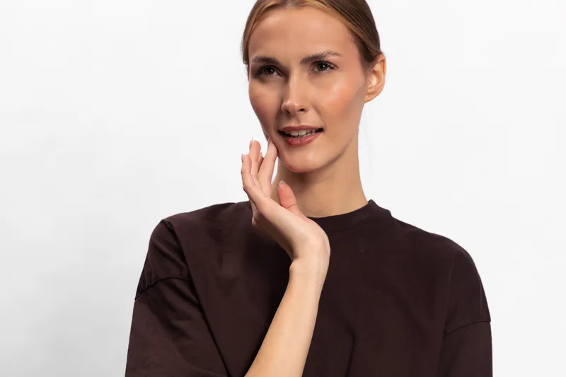 Confident woman with natural makeup and slicked back hair poses with hand near face wearing dark brown top on white background. Concept of beauty, portrait, skincare, and fashion style.