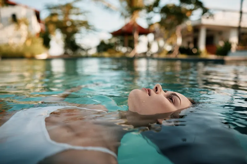 Relaxed woman floats in a clear pool on a sunny day, enjoying serene water, gentle breeze, and a carefree mood, with a calm outdoor resort vibe and focus on wellness and leisure.