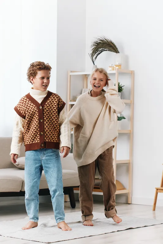 Two cheerful children stand indoors wearing cozy knitted sweaters, barefoot on a light rug, smiling and sharing warmth, capturing candid friendship and playful home comfort.