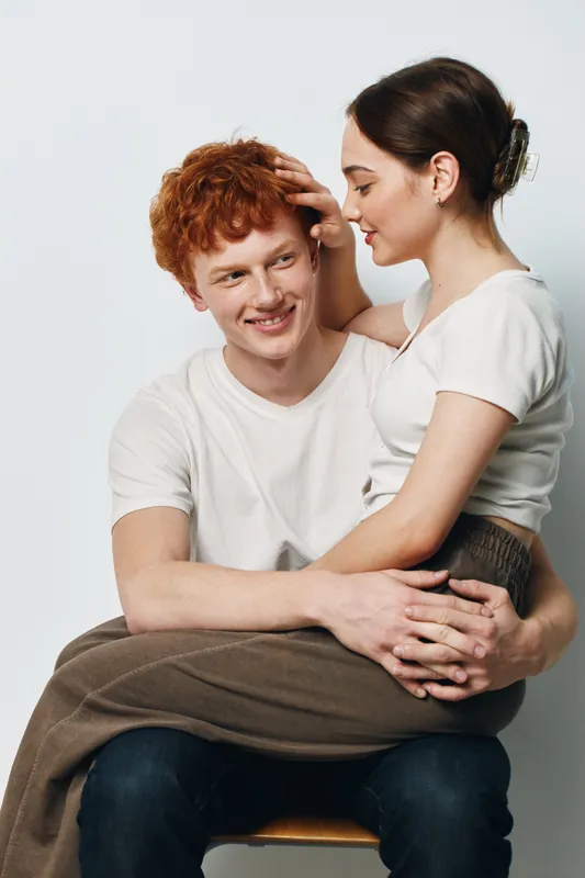 Couple embracing and smiling with affection in casual clothes on neutral background, young woman sitting on man’s lap, enjoying intimate moment and closeness together.