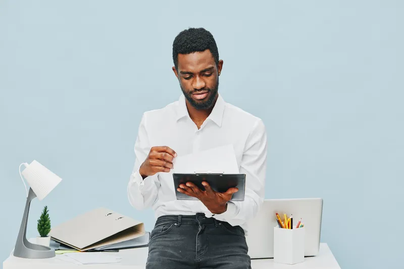 Professional young African American man dressed in white shirt standing at desk and working on tablet device with focused expression, modern workspace, clean background, digital technology concept, wo