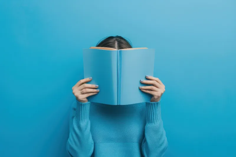 Женщина в синем свитере с книгой, закрывающей лицо, на синем фоне — литературное бегство в красоту и искусство