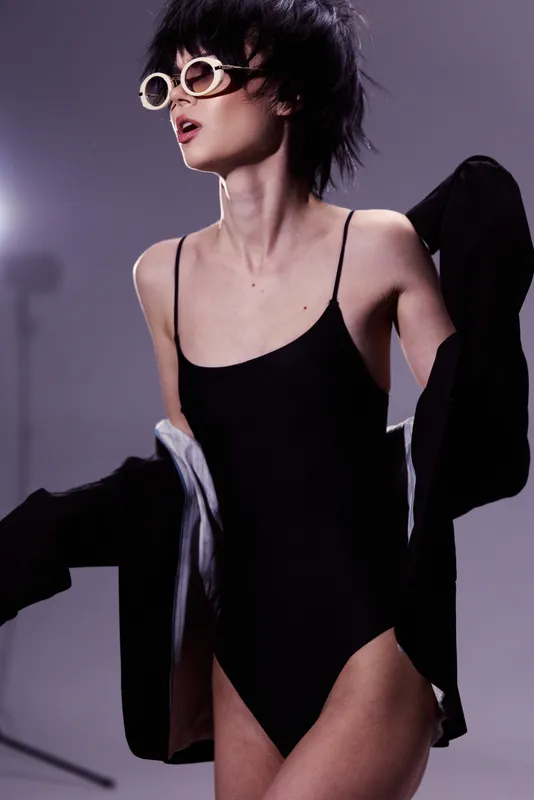 Stylish woman wearing black swimsuit and oversized sunglasses posing indoors with a dark jacket in a modern setting.