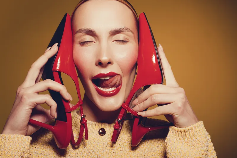 Young woman with closed eyes holds red high heels near face, tongue out playfully. Warm lighting and beige background create a fun and quirky fashion concept.