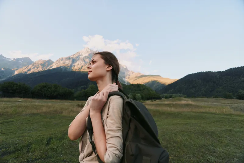 Young woman hiker with backpack stands in an open field, gazing toward mountains, enjoying nature, outdoor travel mood, peaceful moment in golden hour light