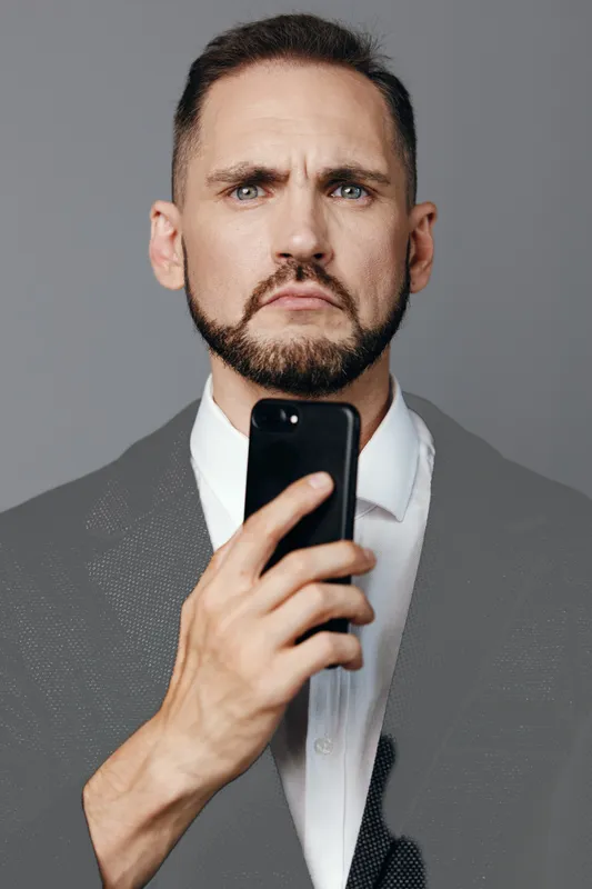 A business professional man stands against a bold colored background, holding a smartphone to convey focus, determination, and a thoughtful, calm mood.