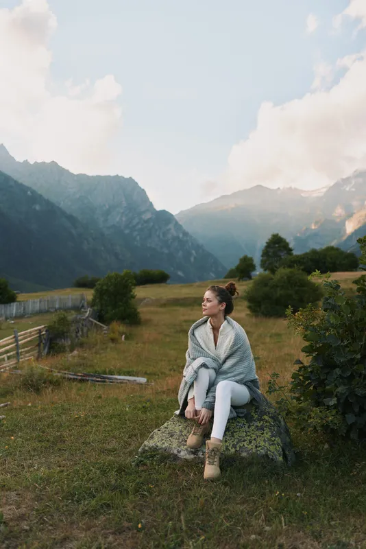 A young woman wrapped in a blanket sits on a rock in a mountainous landscape during a calm day, enjoying the peaceful natural surroundings and fresh air.