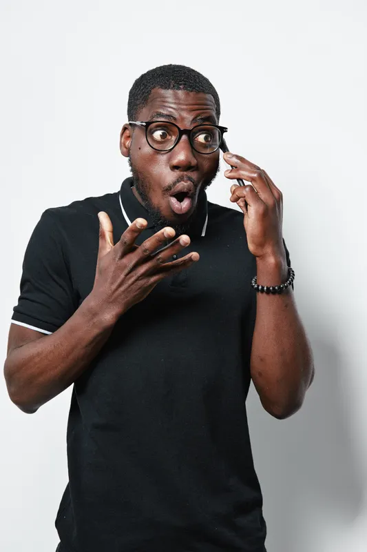 African American man with dark skin and glasses talking on phone with surprised expression, wearing black polo shirt and bracelet on white background.