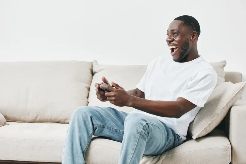 Happy young Black man playing video games on a comfortable couch, expressing joy and excitement in a relaxed setting with a modern aesthetic