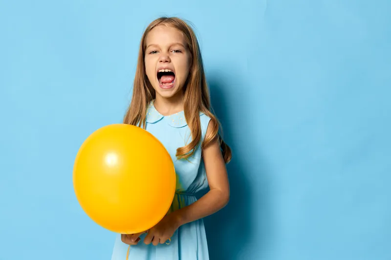 Маленькая девочка в синем платье радостно держит ярко-жёлтый воздушный шар на фоне насыщенного синего фона