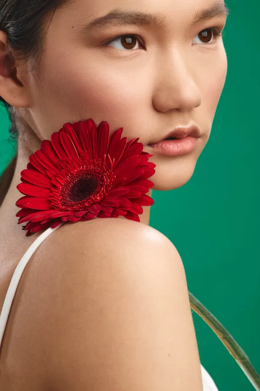 Asian woman gazes softly at the camera with a red gerbera held near her shoulder, conveying beauty and poise in a vibrant studio setting.