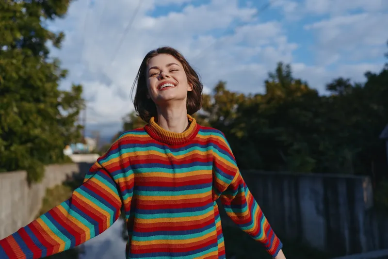 Woman smiling in colorful striped sweater, young adult with short hair, arms out in suburban outdoor scene, casual outfit, joyful expression, lifestyle portrait for travel and fashion