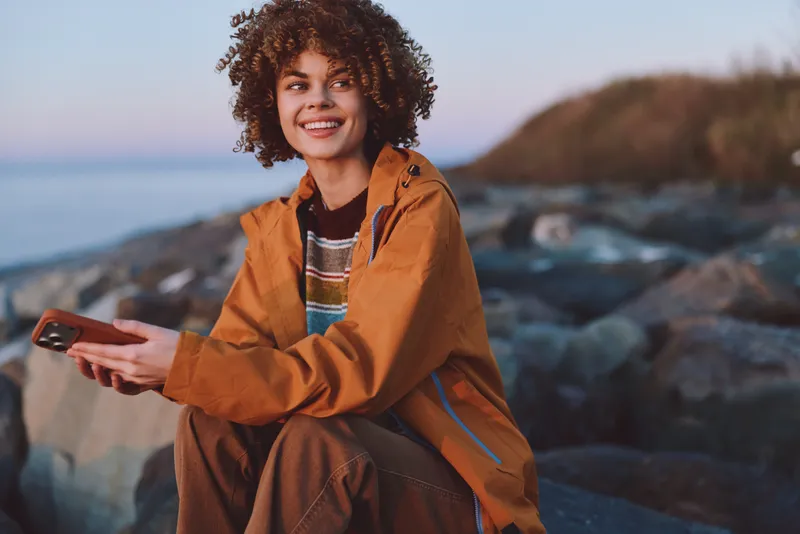 Woman lifestyle portrait on seaside rocks, smiling with curly hair, wearing an orange jacket, holding a smartphone in her hand, relaxed outdoor leisure by the ocean under daylight