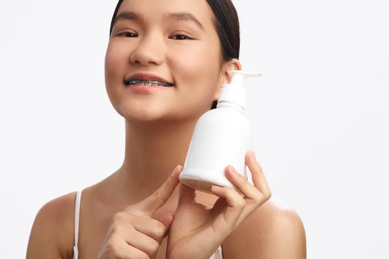 Asian woman with a gentle smile models skincare product in a clean studio, presenting a fresh and confident vibe. She holds a bottle, suggesting a modern beauty routine and everyday care.