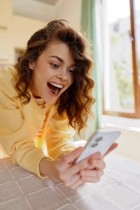 Young woman with curly hair smiles at a smartphone while lying on a bed in a bright home setting, showcasing a casual lifestyle, relaxed moment, and cheerful mood.