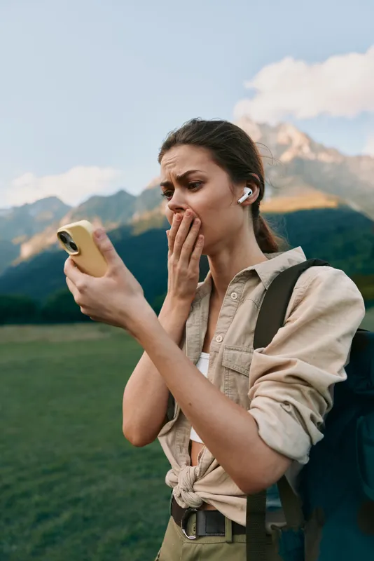 Young woman with earbuds looks shocked while holding a smartphone outdoors with mountains in the background during a clear day.