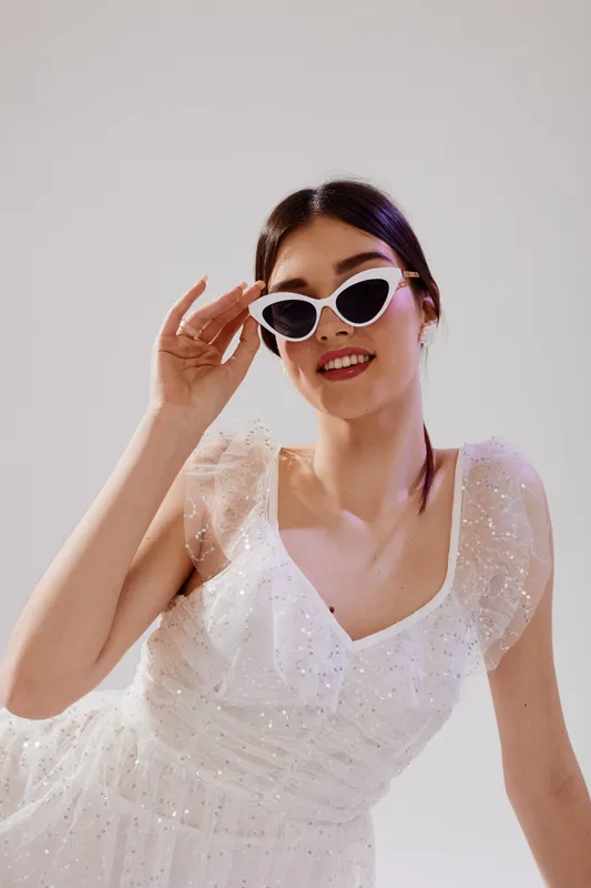 Young woman in a white sparkling dress wearing white cat-eye sunglasses smiles confidently in a studio. Concept of woman beauty fashion, elegance, and modern style.