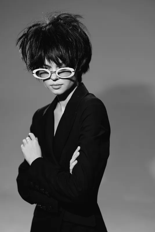 Black and white portrait of a young woman wearing stylish sunglasses and a dark blazer with a modern hairstyle.