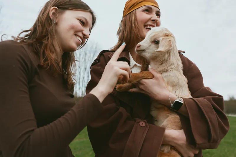 Cheerful woman outdoors, smiling with goat kid, warm moment, casual wear, gentle breeze, natural colors, playful atmosphere, friendship in nature