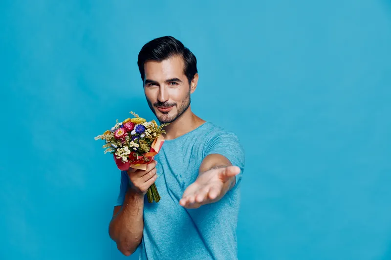 Smiling man in casual blue t-shirt holding colorful bouquet of flowers, reaching out with outstretched hand, isolated on solid turquoise background. Lifestyle concept.
