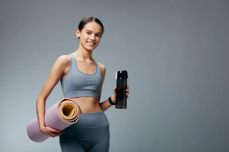 fitness, woman, sport, yoga mat, fitness woman in workout wear holds a rolled yoga mat and a water bottle, ready for training. Clean studio backdrop highlights athletic lifestyle and wellness.