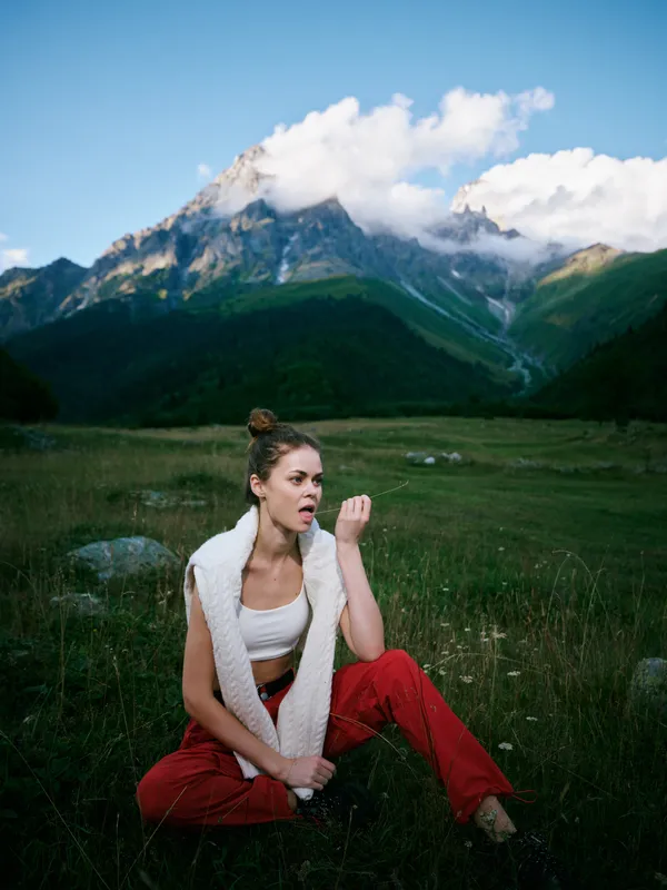 Woman sits in a meadow dressed in red pants and a white scarf, gazing surprised at rugged mountains under a blue sky, capturing outdoor lifestyle, nature and alpine scenery