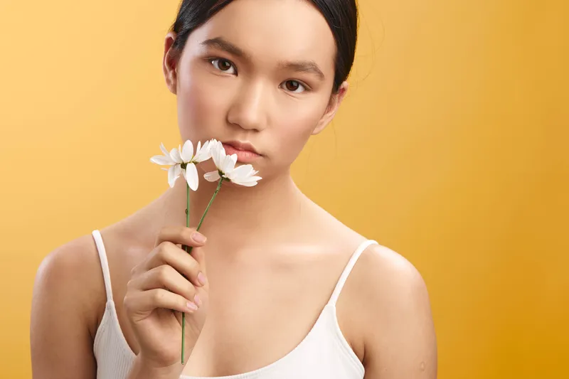 Asian woman softly smiles while holding a white flower near her face against a warm orange backdrop, conveying calm elegance and gentle beauty in a studio portrait.