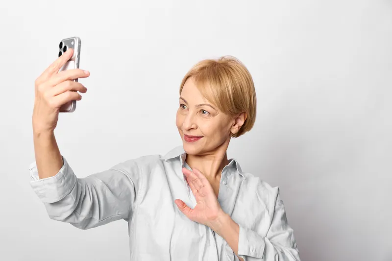 Elderly woman selfie with smartphone, studio portrait on white background, holding phone with one hand and touching chest, softly smiling, surprised expression, copy space