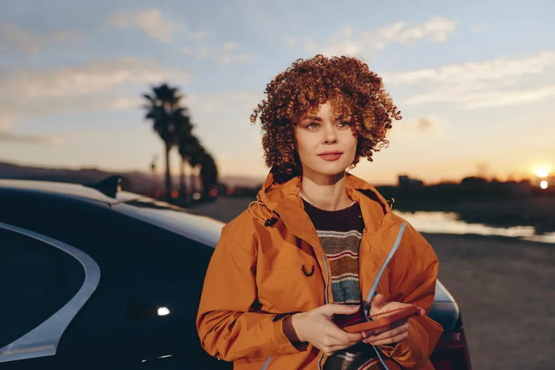 Woman lifestyle sunset car orange jacket curly hair holding phone on parking road near vehicle at dusk, casual travel vibe, looking at camera, natural light, street background