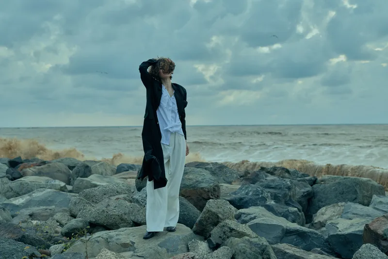 Silhouette on a rocky coast, figure in white pants and dark coat gazing at the windy sea under a cloudy sky, rugged textures and moody lighting