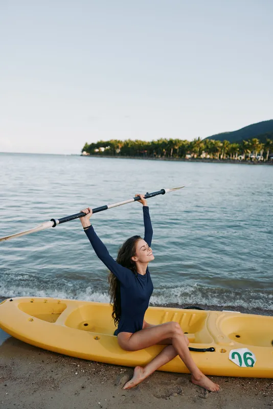 A young woman in a navy swimsuit sits on a yellow kayak by the calm sea, raising a paddle overhead as the shore and hills stretch along the horizon.