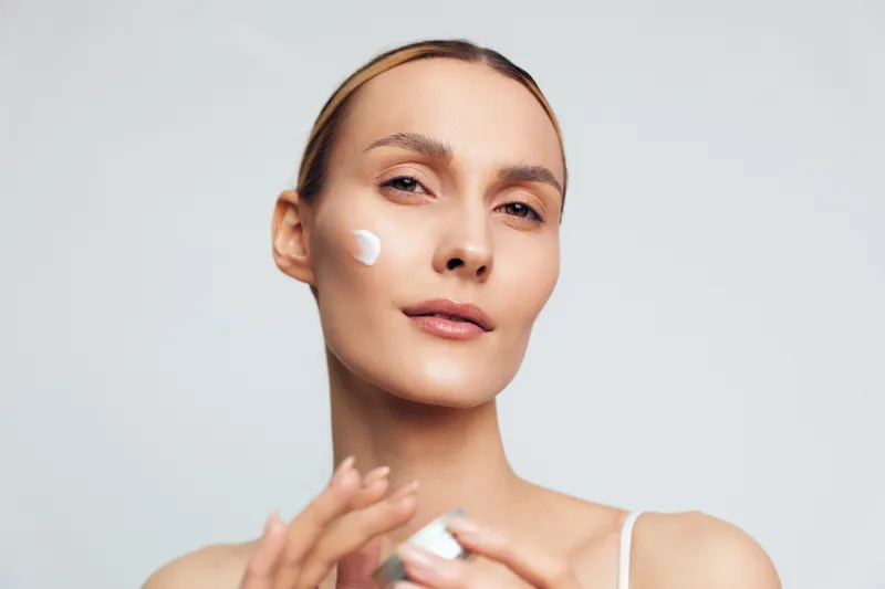 Young woman with clear skin applying white face cream on cheek. Close-up portrait on light background. Concept of skincare, beauty, wellness, and self-care routine. Soft lighting.