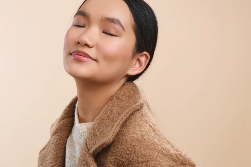 asian woman wearing a cozy coat rests with closed eyes in a soft neutral background, conveying calmness and warmth, yielding a gentle, introspective mood in a simple portrait.