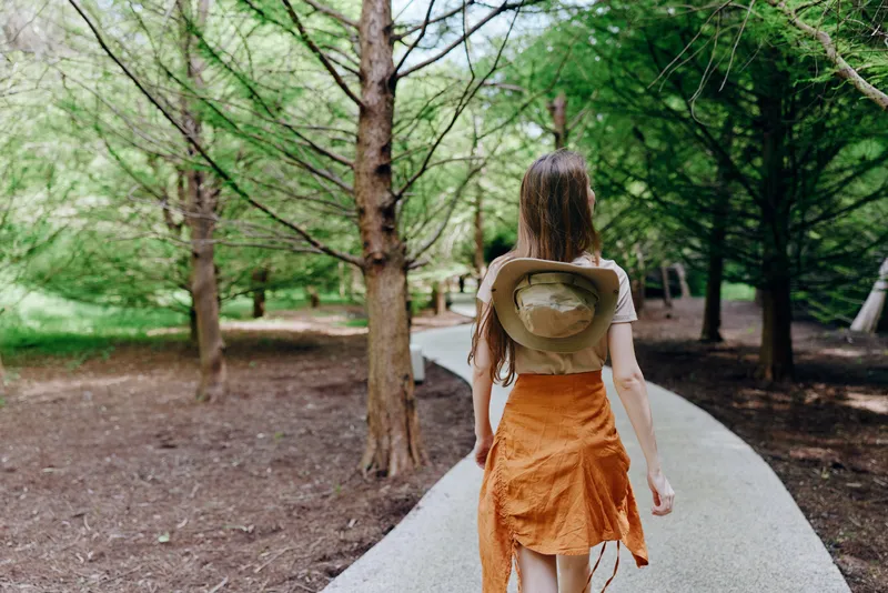Woman walking on a path in a green forest with a straw hat on her back and an orange skirt, summer outdoors scene with trees, trail and a peaceful nature escape.