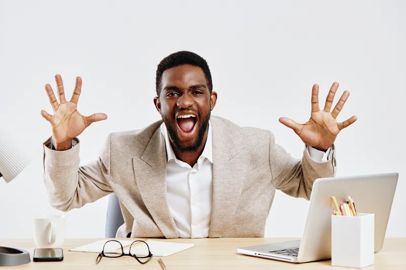 Confident smiling young African American man in beige blazer and white shirt gesturing with hands open, showing excitement and energy, sitting at desk with laptop computer and stationery, studio shot.