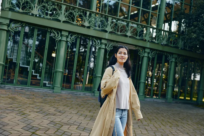 Woman in a beige coat and light sweater walks outdoors beside a glass building, showcasing urban fashion, casual style, and confident streetwear in a sunny city setting.