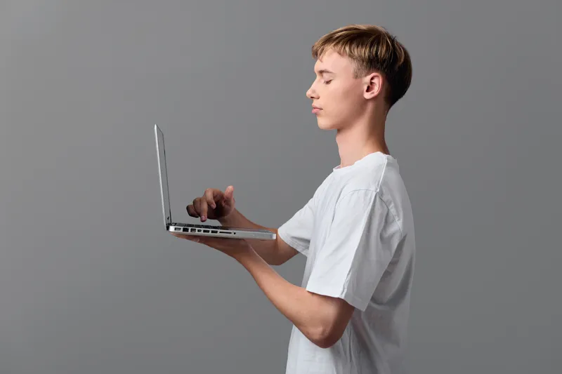 Casual young man typing on a slim laptop held in one hand, focused expression while working, modern technology in a clean studio environment, neutral grey backdrop