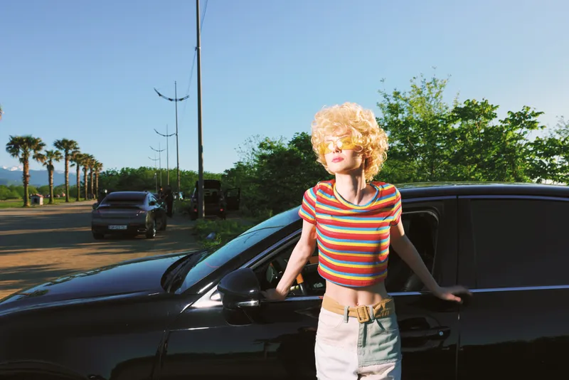 Smiling woman with curly blonde hair in a striped crop top leans on a dark car outdoors, capturing a carefree lifestyle with retro film color and sunny summer mood and relaxed vibe today.