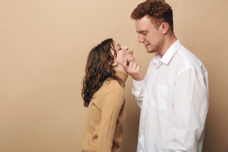 Young couple in casual clothes share a tender moment, man gently touching woman’s chin. Warm beige background creates intimate and loving atmosphere, concept of romance and affection.