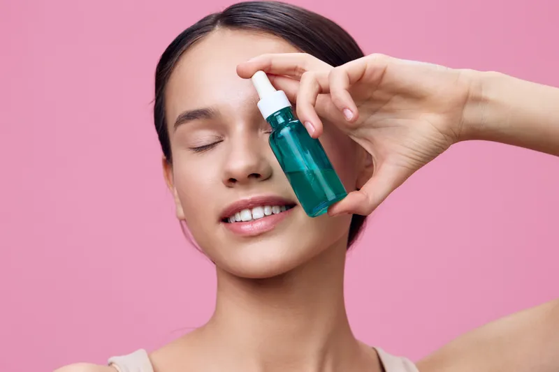 Young woman holding green skincare serum bottle with closed eyes, smiling against a pink backdrop, promoting skincare and beauty