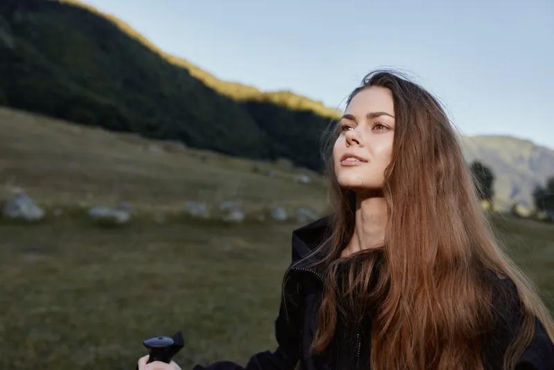 A thoughtful young woman with long hair stands outdoors in a wide open landscape, looking upward with a calm expression, conveying curiosity and serenity in natural light.