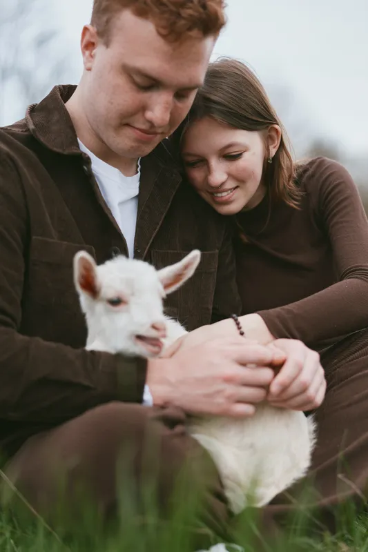 Loving couple outdoors, sharing a gentle moment with a playful goat kid on a grassy meadow, warm natural light, lifestyle portrait emphasizing bonding, affection, and happiness.