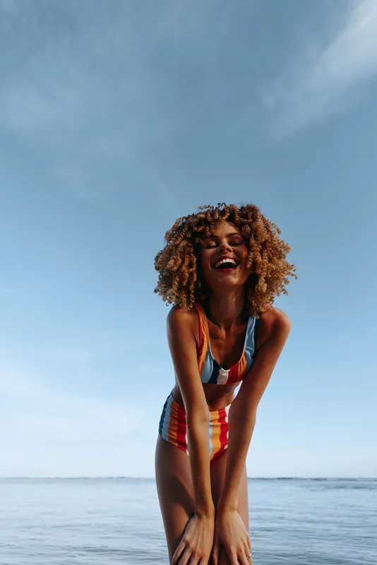 Woman curly hair laughing on beach by ocean, summer outdoor scene under blue sky, close to camera, joyful carefree mood, natural light, vertical portrait, young adult, smiling, seaside relaxation