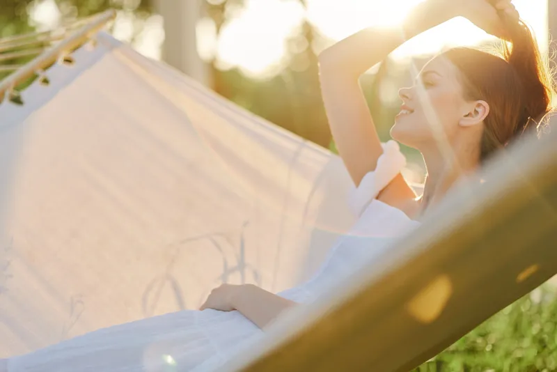 Young woman relaxing in a hammock, wearing a white dress, surrounded by nature, enjoying a sunny day, embodying tranquility and peace