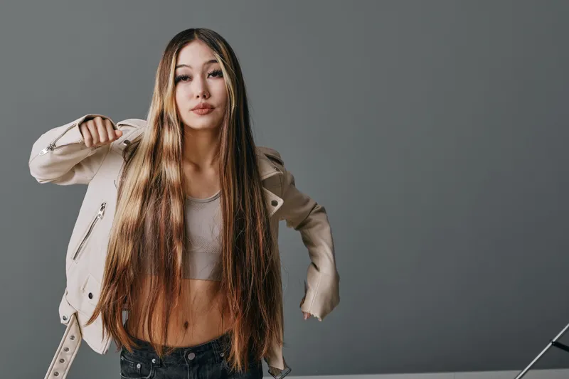 Young woman with long hair wearing jacket and crop top posing against gray background with confident expression in studio for fashion portrait and style concept.