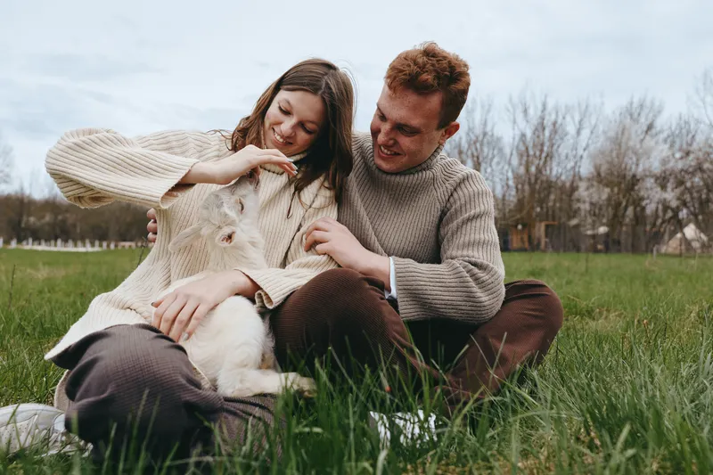 Caring couple outdoors with a goat kid, sharing a tender moment on the grass. Cozy sweaters, natural light, relaxed mood, rustic backdrop. Emphasis on family, animal companionship, and simple
